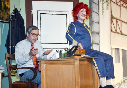 Ein Mann im Anzug sitzt an einem Schreibtisch, während ein clownartiger Charakter daneben lächelt.