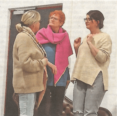 Drei Frauen in modischen Oberteilen unterhalten sich in einem Raum mit einer braunen Couch im Hintergrund.