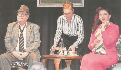 Drei Schauspieler auf der Bühne: ein Mann im Anzug, eine Bedienung mit Tablett und eine Frau in pinker Kleidung.