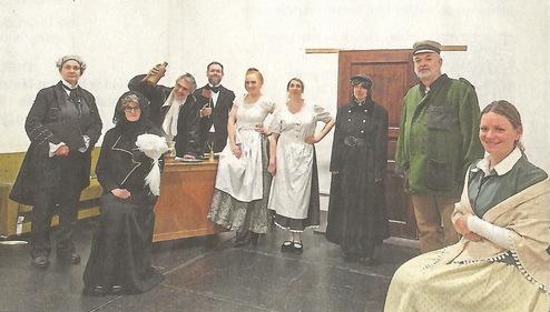 Gruppe von zehn Personen in historischer Kleidung, darunter eine Frau in schwarzem Kleid und Männer in Anzügen, vor einer Wand.