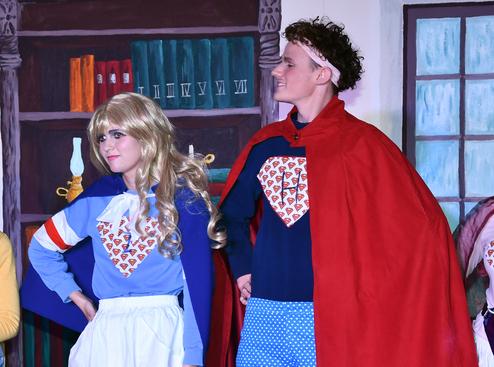 Zwei Personen in Superheldenkostümen stehen nebeneinander auf einer Bühne, umgeben von Bücherregalen.