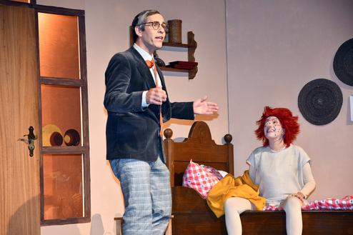 Schauspieler in formeller Kleidung spricht lebhaft zu einer Person mit roter Perücke auf einem Bett, im Hintergrund ein Fenster.