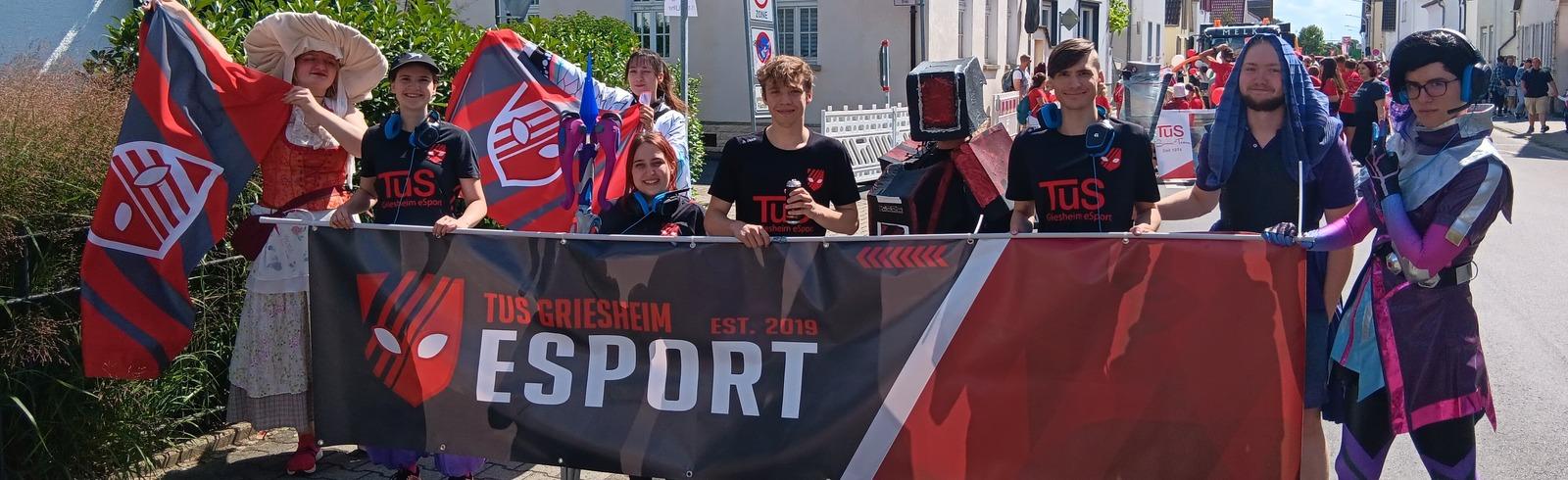 Gruppierung von Menschen, die ein Banner mit &bdquo;TUS Griesheim Esport&ldquo; halten, umgeben von bunten Fahnen in einer Stra&szlig;e.