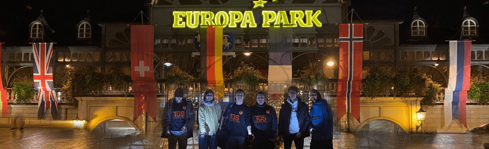 Gruppe von sechs Menschen vor dem beleuchteten Eingang des Europa Parks mit Fahnen im Hintergrund.
