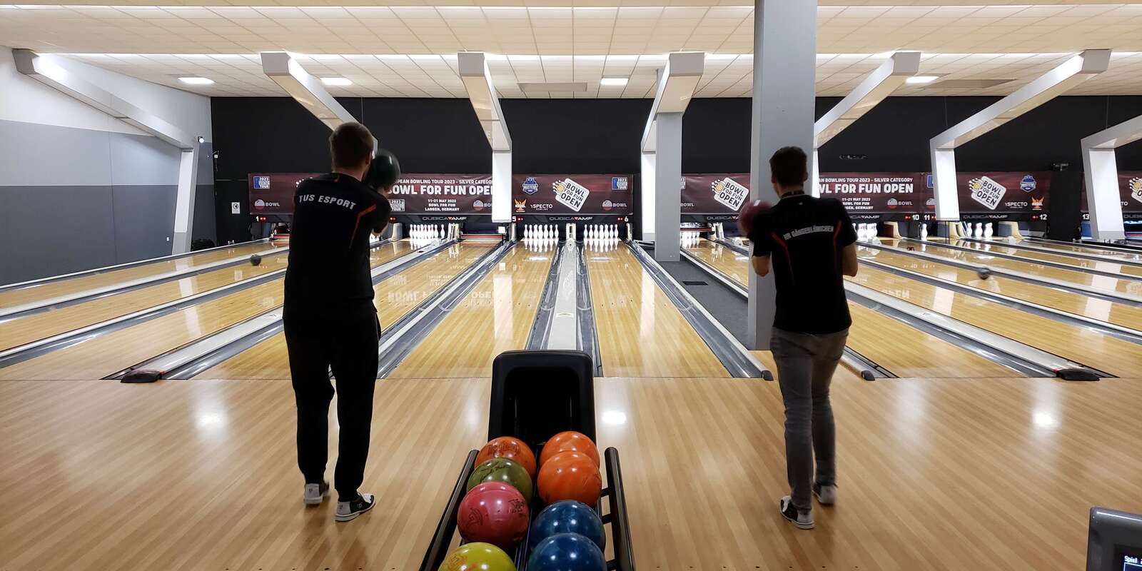 Zwei Spieler in Sportbekleidung werfen Bowlingkugeln auf eine gut beleuchtete Bahn mit mehreren Pins im Hintergrund.