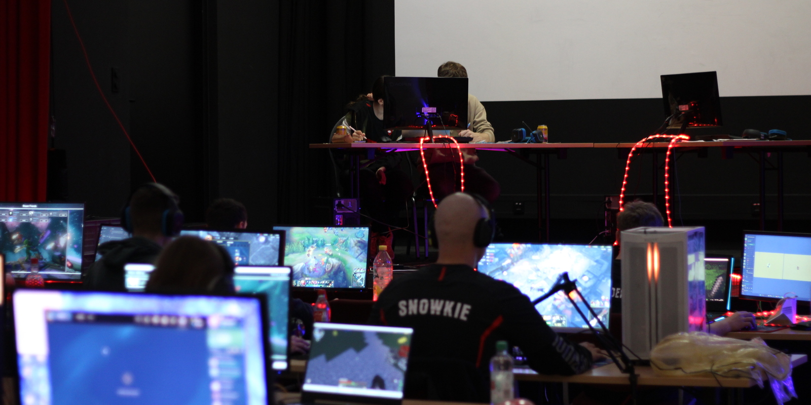 Gruppenszene eines Gaming-Events mit mehreren Spielern an Computern und zwei Personen an einem Tisch im Hintergrund.