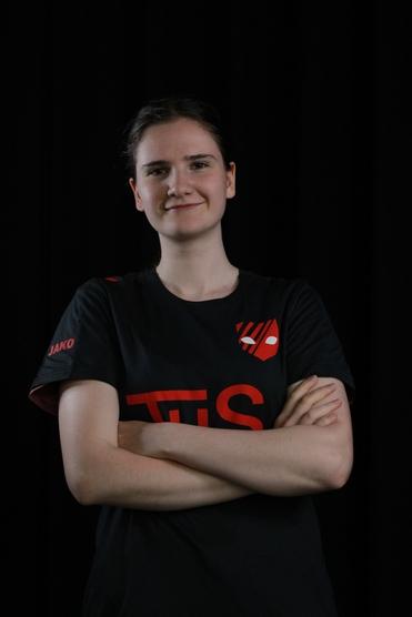 Porträt einer jungen Frau mit verschränkten Armen, die ein schwarzes Shirt mit rotem Logo trägt, vor schwarzem Hintergrund.