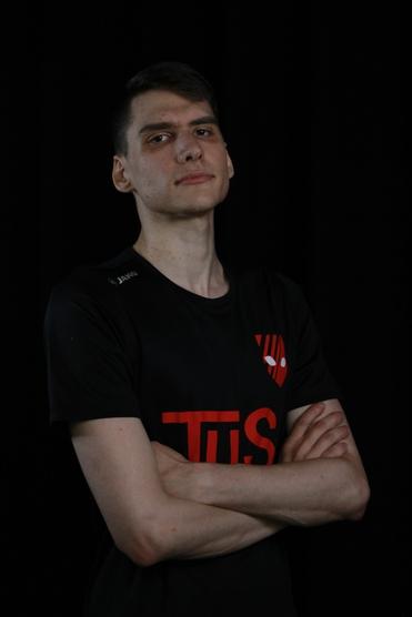 Junger Mann mit verschränkten Armen, trägt ein schwarzes T-Shirt mit rotem Logo auf schwarzem Hintergrund.