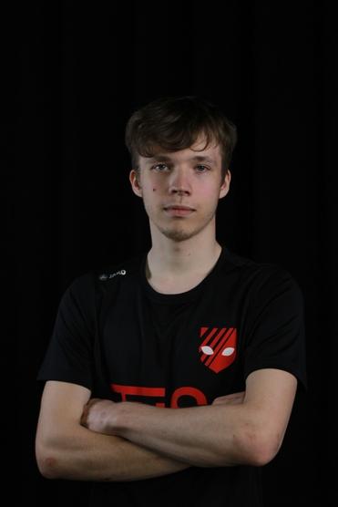 Porträt eines jungen Mannes mit verschränkten Armen in einem schwarzen T-Shirt mit rotem Logo, vor schwarzem Hintergrund.