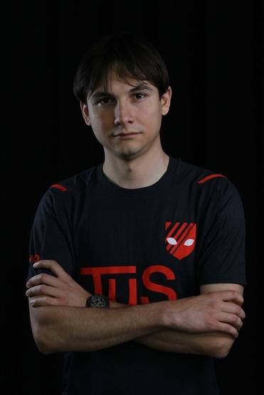 Porträt eines Mannes mit crossed arms, getragenem schwarzem T-Shirt mit rotem Logo und neutralem Gesichtsausdruck.