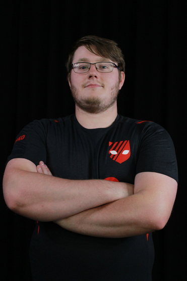 Junger Mann mit Brille, Arme verschränkt, trägt ein schwarzes Shirt mit rotem Logo vor schwarzem Hintergrund.