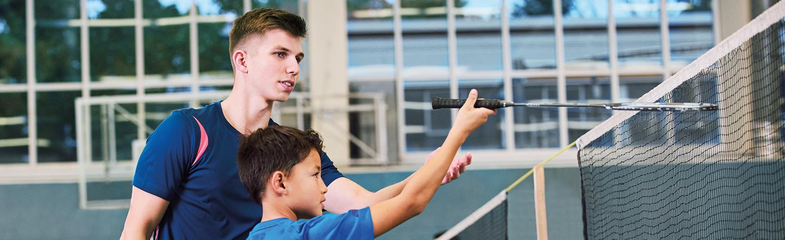 Ein Trainer zeigt einem Jungen mit dem Shirt &bdquo;DETERS GERMANY&ldquo; die richtige Technik am Badminton-Netz.