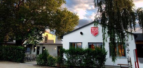 Ein sportliches Vereinsgebäude mit einem großen Baum und Tischgarnitur im Freien, beschattet von einer Wolkendecke.