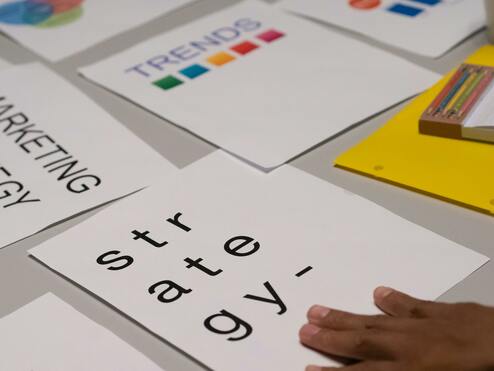 Papiere mit den Worten „STRATEGY“ und „MARKETING STRATEGY“ liegen auf einem Tisch, daneben Hand und ein Block mit Stiften.