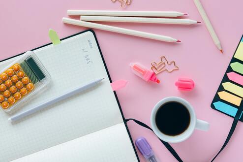 Schreibtisch-Zubeh&ouml;r: Notizbuch, Bleistift, Marker, Kaffeetasse und orangefarbene Kugelschreiber auf pinkem Hintergrund.