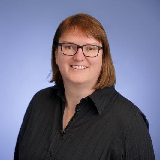 Portrait einer lächelnden Person mit braunen Haaren, die eine schwarze Bluse trägt, vor einem blauen Hintergrund.