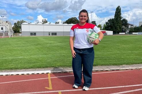 Junge Frau in Sportoutfit hält einen Fußball, steht auf einem Rasenplatz mit Tartanbahn und Sporteinrichtungen im Hintergrund.