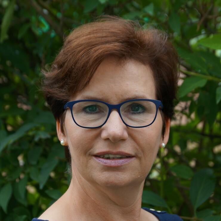 Porträt einer Frau mit kurzen braunen Haaren, blauen Brillen und gestreiftem Oberteil, im Freien vor grünen Pflanzen.