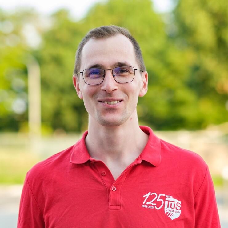 Portrait eines Mannes in rotem Poloshirt mit Logo, lächelnd vor unscharfem grünen Hintergrund.