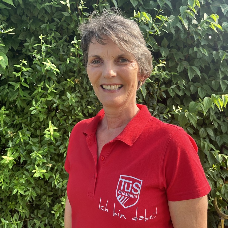 Frau mit kurz geschnittenem Haar lächelt, trägt ein rotes Poloshirt mit Aufdruck vor grünem Hintergrund.