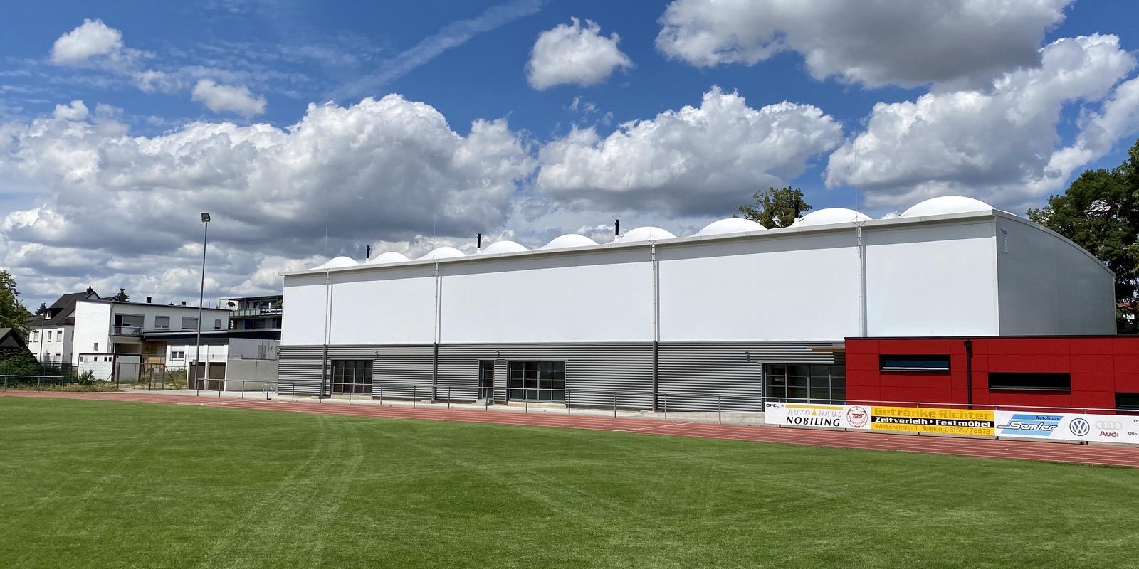 Moderne Sporthalle mit grauer Fassade und roten Akzenten, umgeben von grünem Rasen und wolkenlosem Himmel.