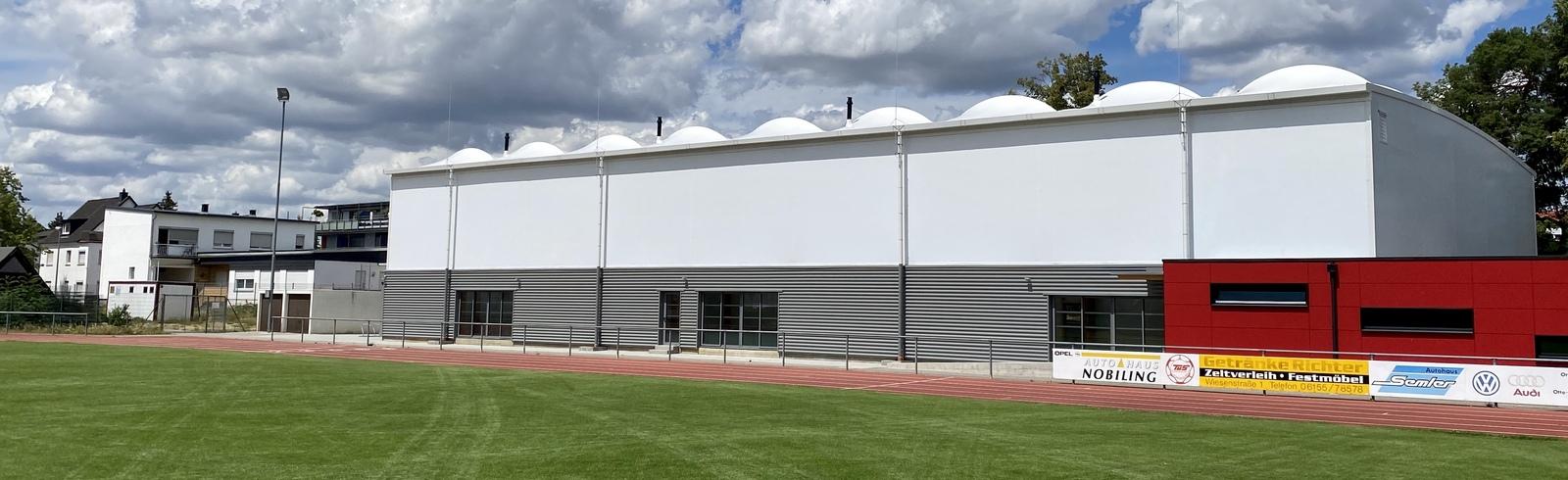 Moderne Sporthalle mit grauer Fassade und roten Akzenten, umgeben von gr&uuml;nem Rasen und wolkenlosem Himmel.