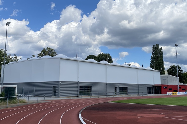 Modernes Sportgebäude mit gewölbtem Dach, umgeben von Laufbahn und grünem Rasen, unter blauem Himmel mit Wolken.