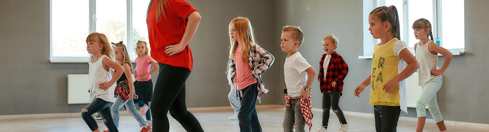 Tanzende Kinder in einem Trainingsraum, angeleitet von einer Frau, kreative Bewegungen zur Musik.