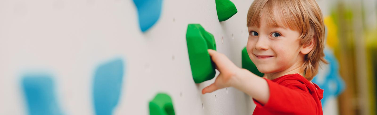 Junge mit blonden Haaren klettert an einer bunten Kletterwand mit gr&uuml;nen und blauen Griffen.