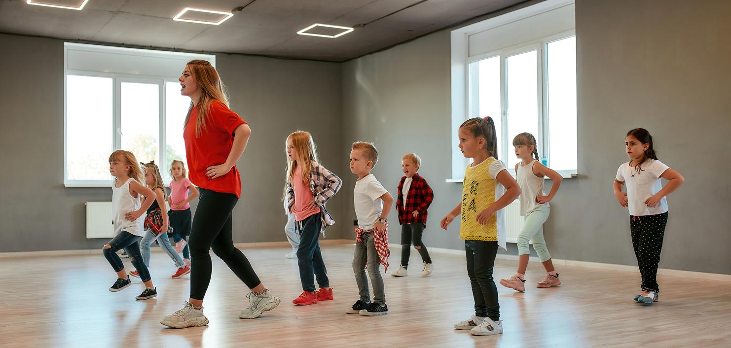 Tanzende Kinder in einem Trainingsraum, angeleitet von einer Frau, kreative Bewegungen zur Musik.