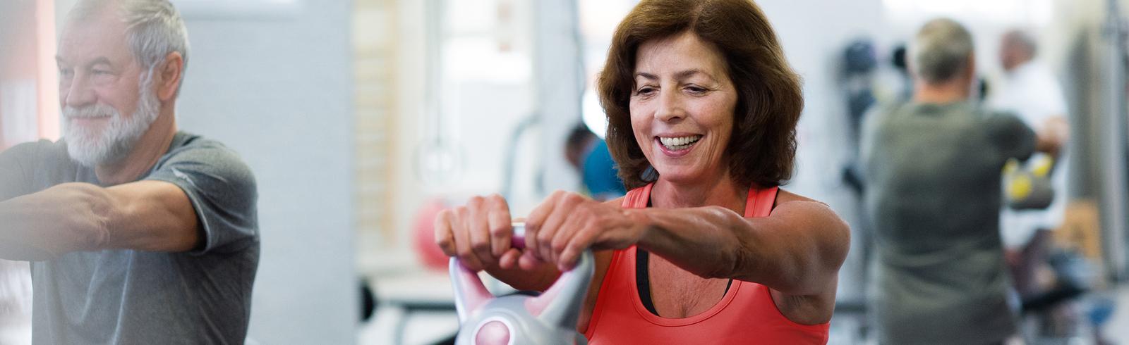 &Auml;ltere Frau in Sportkleidung hebt eine Kettlebell im Fitnessstudio, l&auml;chelt dabei und trainiert.
