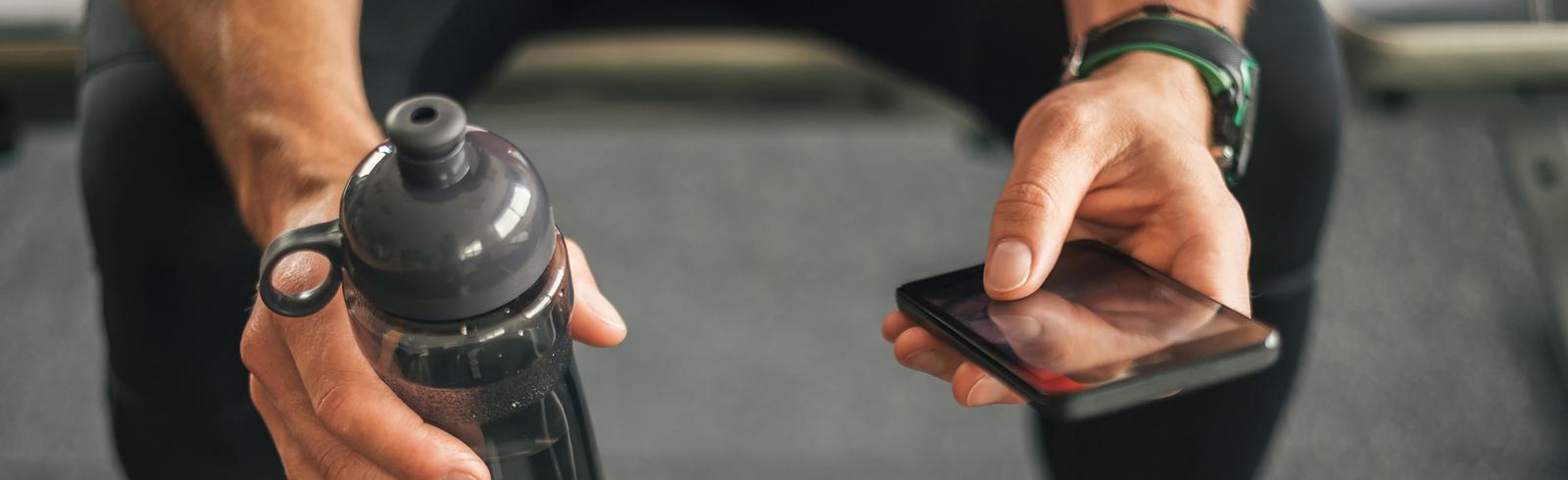 Mann sitzt auf einem Fitnessgerät und hält eine Wasserflasche und ein Smartphone in den Händen.
