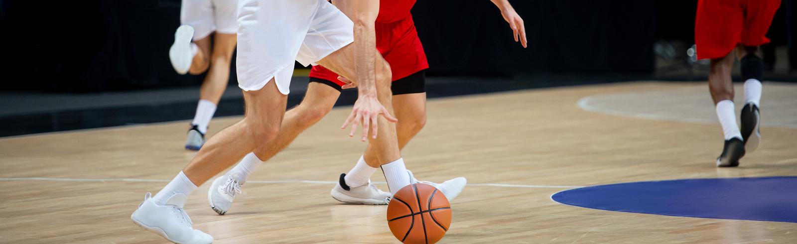 Spielende Basketballer auf einem Spielfeld, w&auml;hrend ein Ball zwischen ihren F&uuml;&szlig;en rollt.