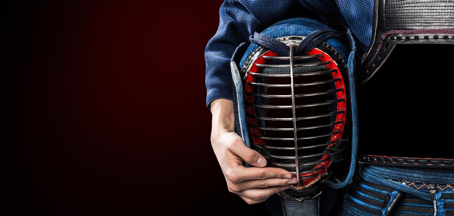Detailansicht eines Kendo-Praktizierenden mit einer rot-schwarzen Men-Doppelmaske auf dem Arm, in traditioneller Uniform.