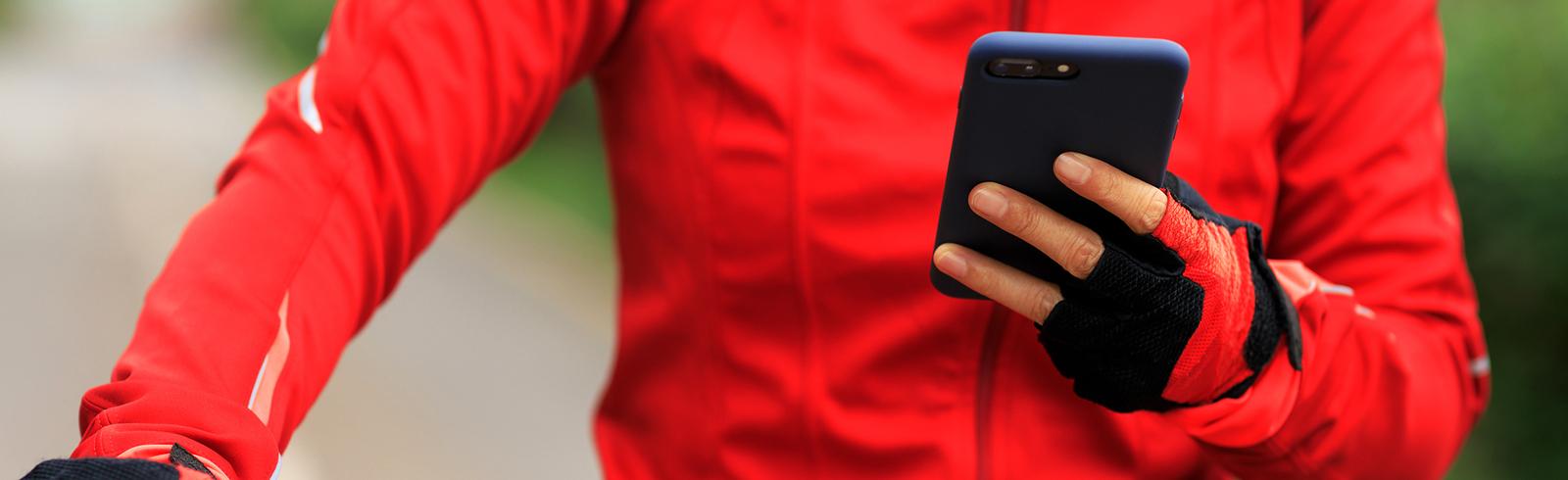 Person in roter Jacke h&auml;lt ein Smartphone und greift mit der anderen Hand den Lenker eines Fahrrads auf einem Weg.