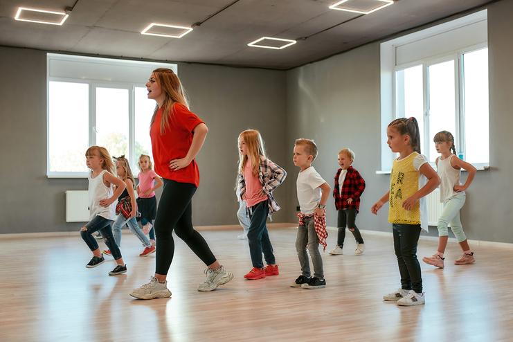 Tanzende Kinder in einem Trainingsraum, angeleitet von einer Frau, kreative Bewegungen zur Musik.