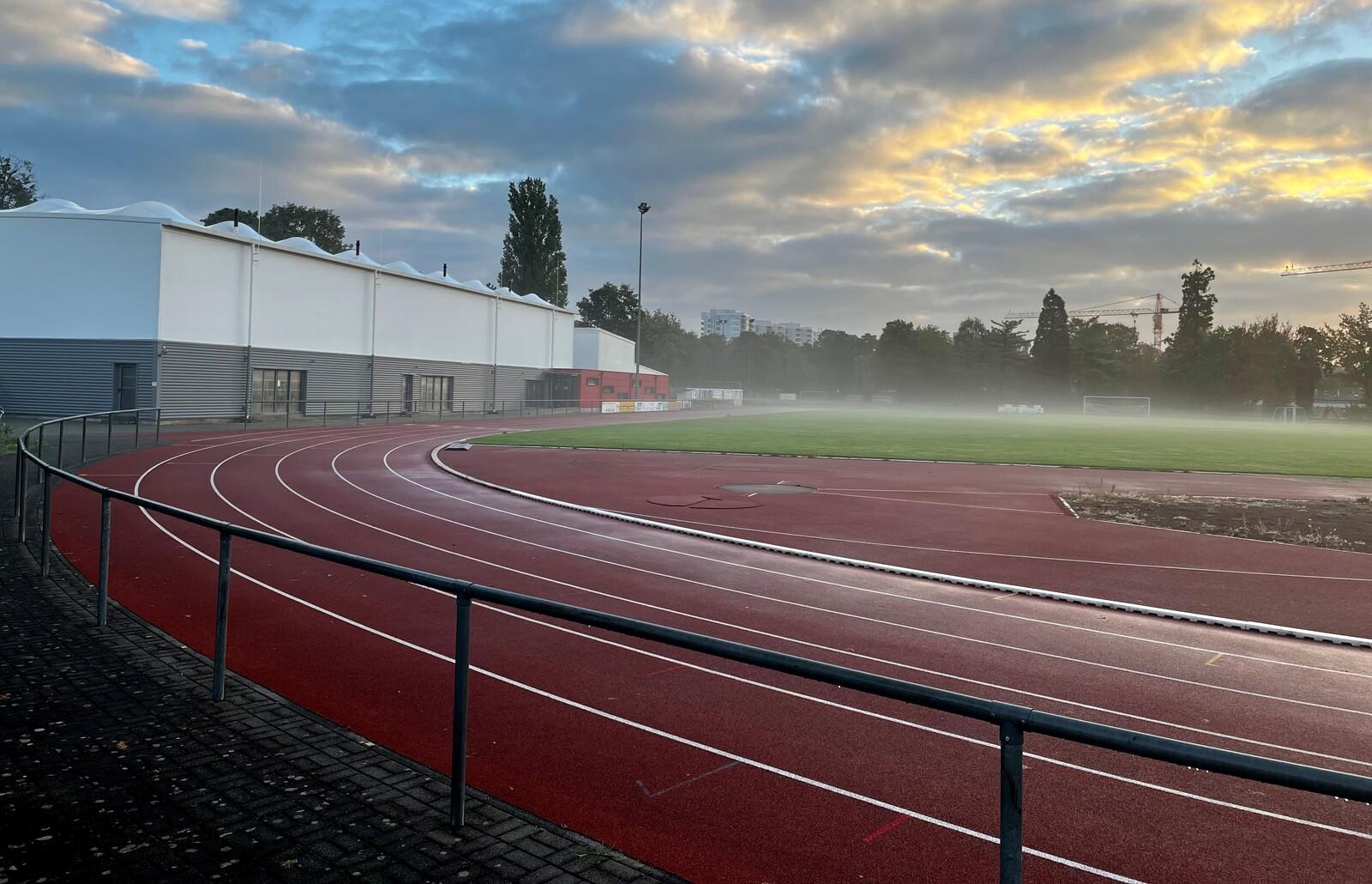 Sportplatz mit Laufbahn, verschwommener Rasenfl&auml;che und einem modernen Geb&auml;ude im Hintergrund bei nebligem Wetter.