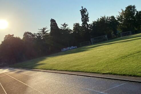 Sonnenaufgang &uuml;ber einem gr&uuml;nen Fu&szlig;ballfeld mit Toren, umgeben von B&auml;umen und einer Laufbahn im Vordergrund.