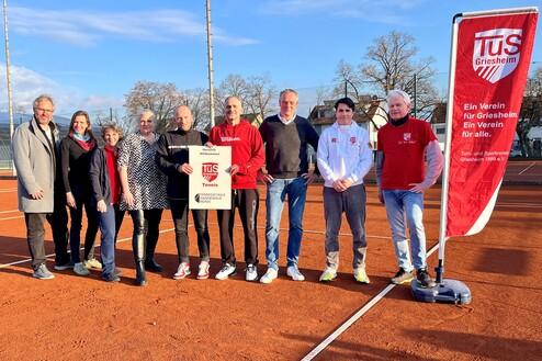 Gruppierung von neun Personen auf einem Tennisplatz, mit einem Banner des TuS Griesheim im Hintergrund.