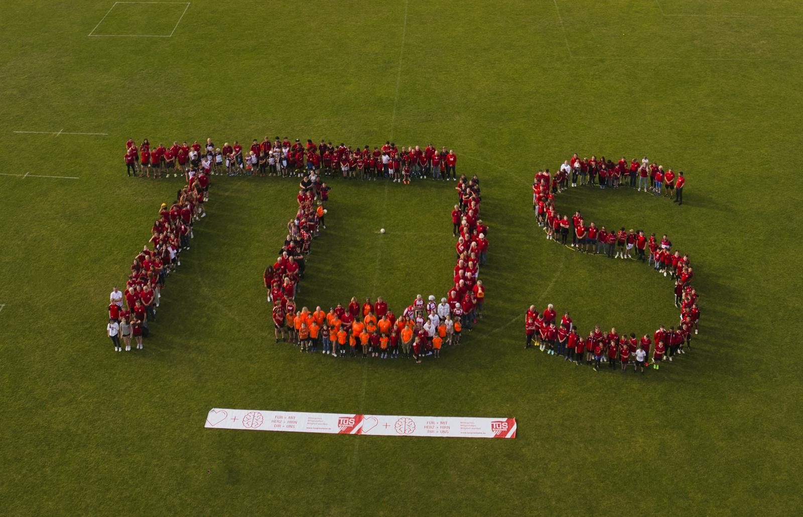 Gruppierung von Menschen in rot und orange bildet den Schriftzug "TDS" auf einem gr&uuml;nen Sportfeld unter blauem Himmel.