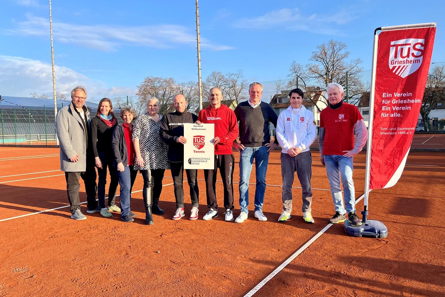 Gruppierung von neun Personen auf einem Tennisplatz, mit einem Banner des TuS Griesheim im Hintergrund.