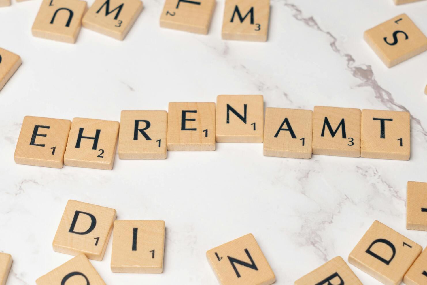 Wortspiel mit Holzbuchstaben auf Marmoroberfläche, das das Wort "EHRENAMT" bildet. Andere Buchstaben sind verstreut.