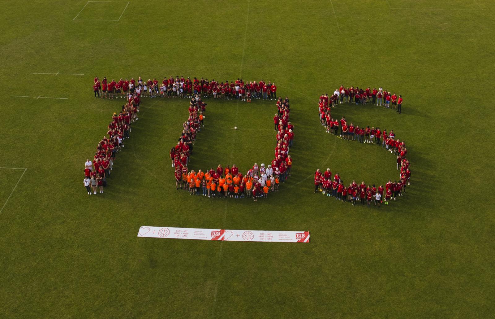Gruppierung von Menschen in rot und orange bildet den Schriftzug "TDS" auf einem gr&uuml;nen Sportfeld unter blauem Himmel.
