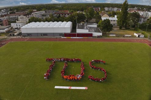 Gruppierung von Menschen in rot und orange bildet den Schriftzug "TDS" auf einem grünen Sportfeld unter blauem Himmel.