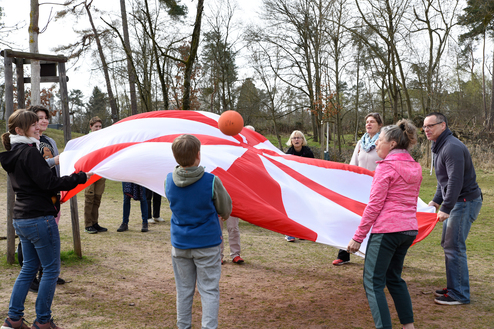 Gruppe von Menschen spielt mit einem großen, bunten Fallschirm und einem Ball auf einer Wiese im Freien.