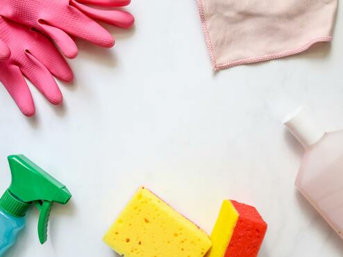 Reiniger, gelber und roter Schwamm, pinke Handschuhe und ein Spr&uuml;hflasche auf einer wei&szlig;en Oberfl&auml;che arrangiert.
