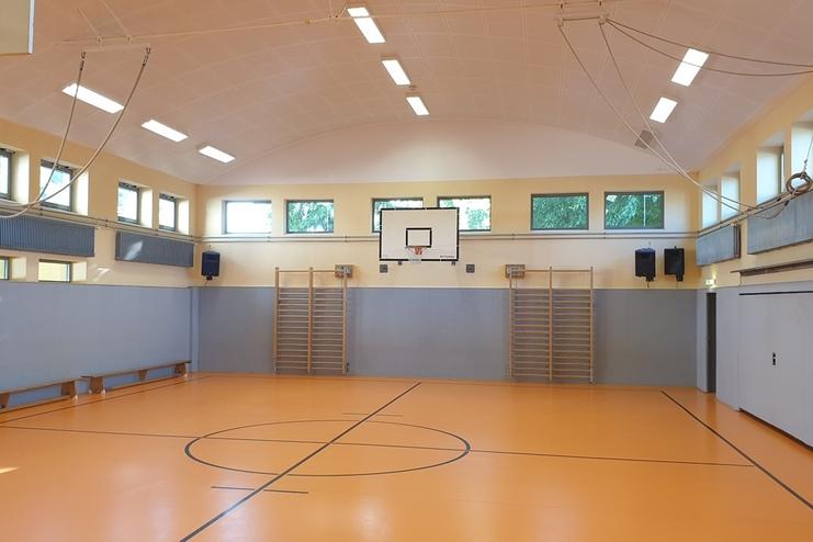 Blick in eine Sporthalle mit orangem Boden, Basketballkorb und mehreren W&auml;nden mit Turnger&auml;ten und Fenstern.
