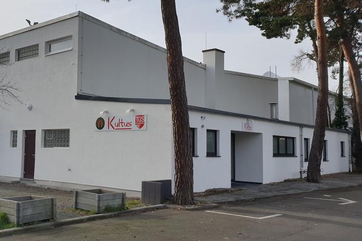 Eingangsbereich eines wei&szlig;en Geb&auml;udes mit dem Schriftzug &bdquo;Kultus&ldquo; und parkendem blauen Auto im Vordergrund.