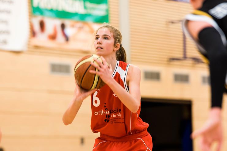 Basketballspielerin in rot, bereit zum Wurf, hält einen Ball in der Halle mit Werbebannern im Hintergrund.