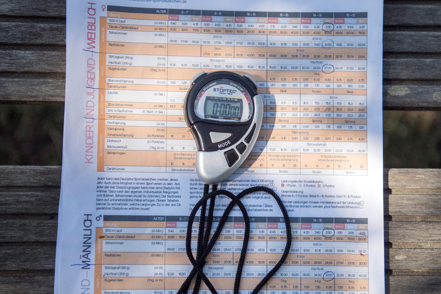 Stoppuhr und Infoblatt des Deutschen Sportabzeichens auf einer Holzbank im Freien.
